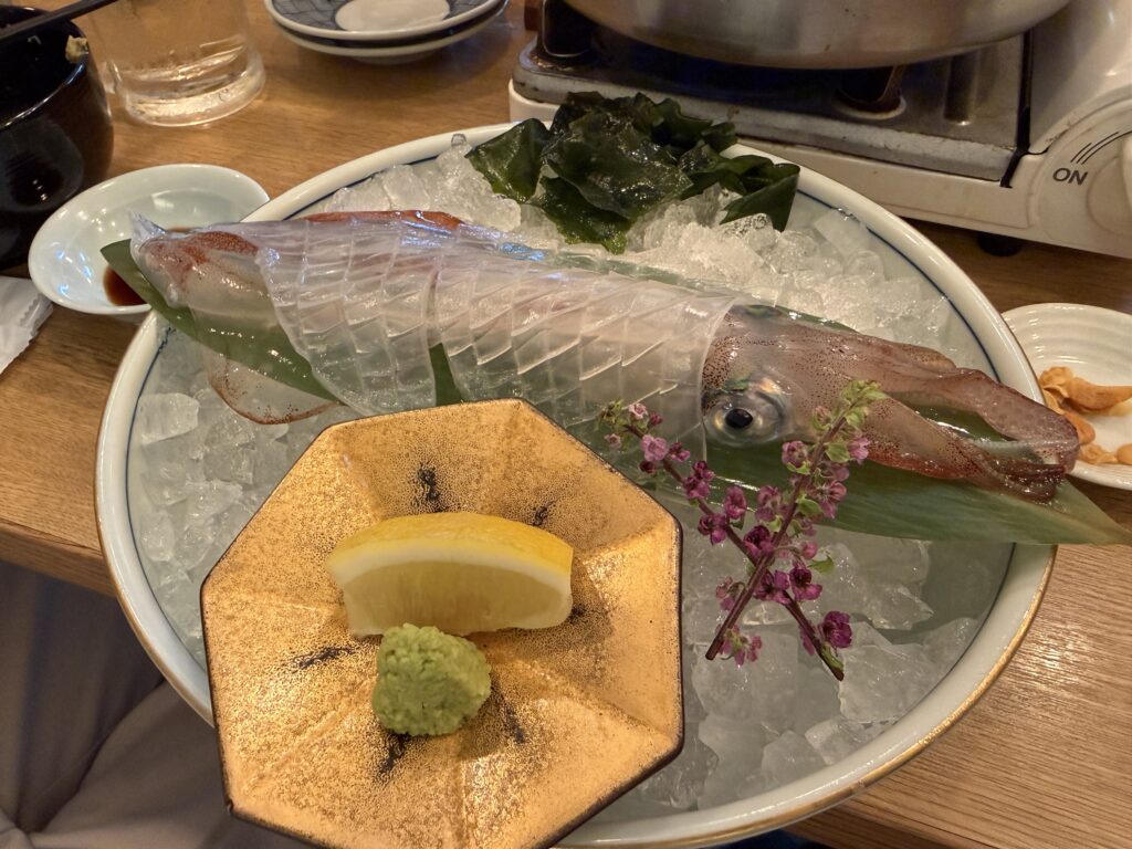 捌きたて活きイカの姿造りを上から捉えた一皿で、透き通った身と整然と並ぶ細い切り込みが美しく、博多もつ鍋の店で味噌仕立ての鍋と一緒に味わった福岡ならではのコリコリ食感と甘みを伝える名物料理