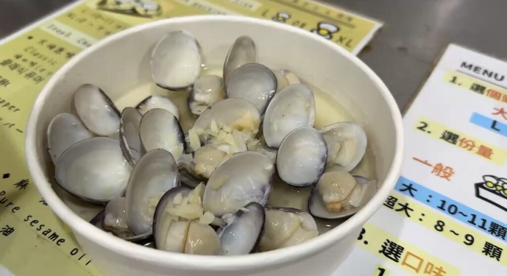 饒河街観光夜市の屋台で味わったガーリック風味の蒸し蛤を間近で捉えた一皿で、刻みニンニクと香味だれがたっぷり絡んだ貝の様子から台北夜市らしい屋台グルメの魅力が伝わるカット