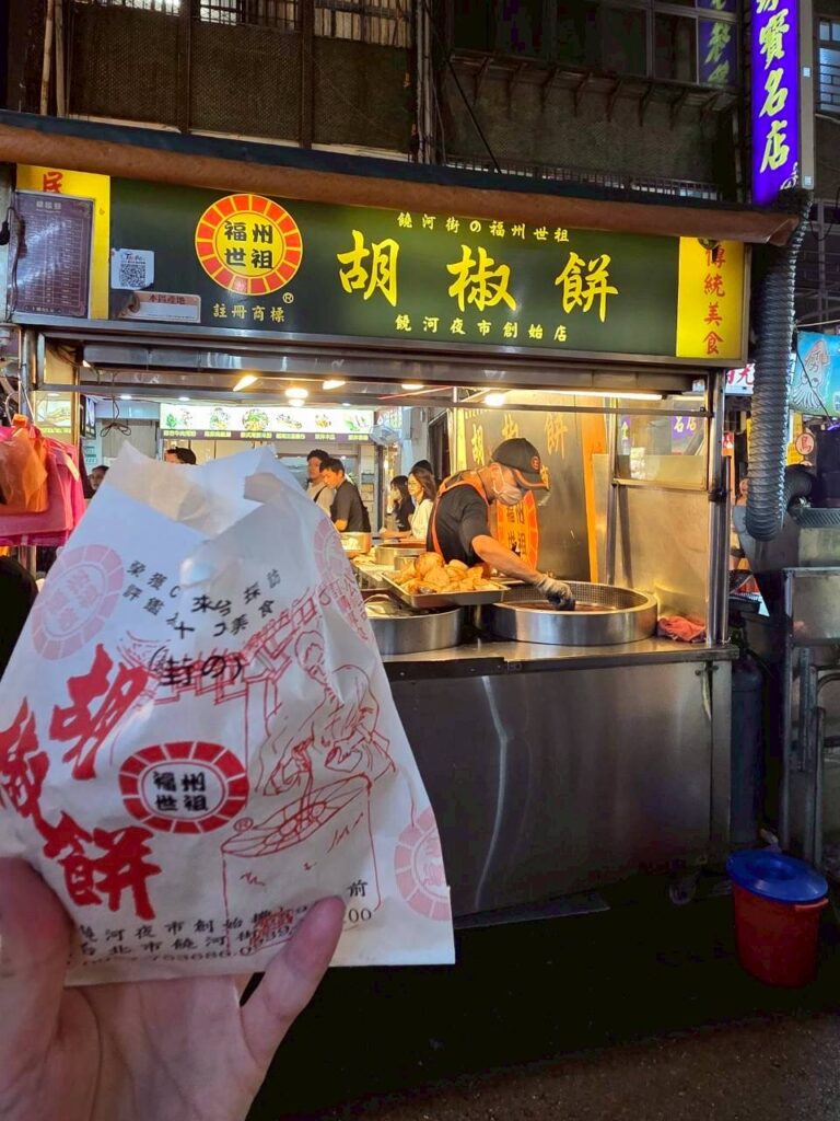 台北・饒河街観光夜市で名物胡椒餅を販売する屋台前に長く伸びる行列の様子を捉えた縦長スナップ。夜の賑わいの中、回転が速く待ち時間が比較的短かった人気店の活気と、3つの夜市の中で最も混雑していた饒河街の雰囲気が伝わる一枚