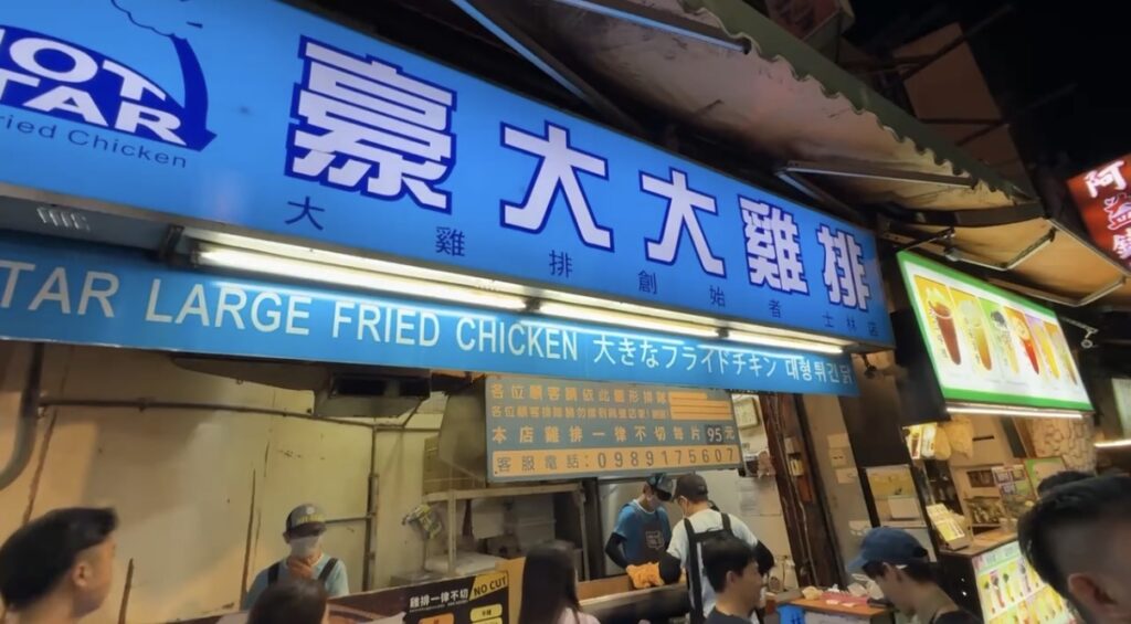 台北・士林夜市で立ち寄った名物グルメ「大鶏排」の屋台を捉えたスナップ。揚げたての大きなフライドチキンが並ぶ店頭の様子から、食べ歩きとゲームの両方を楽しめる広い夜市の活気ある雰囲気が伝わる一枚